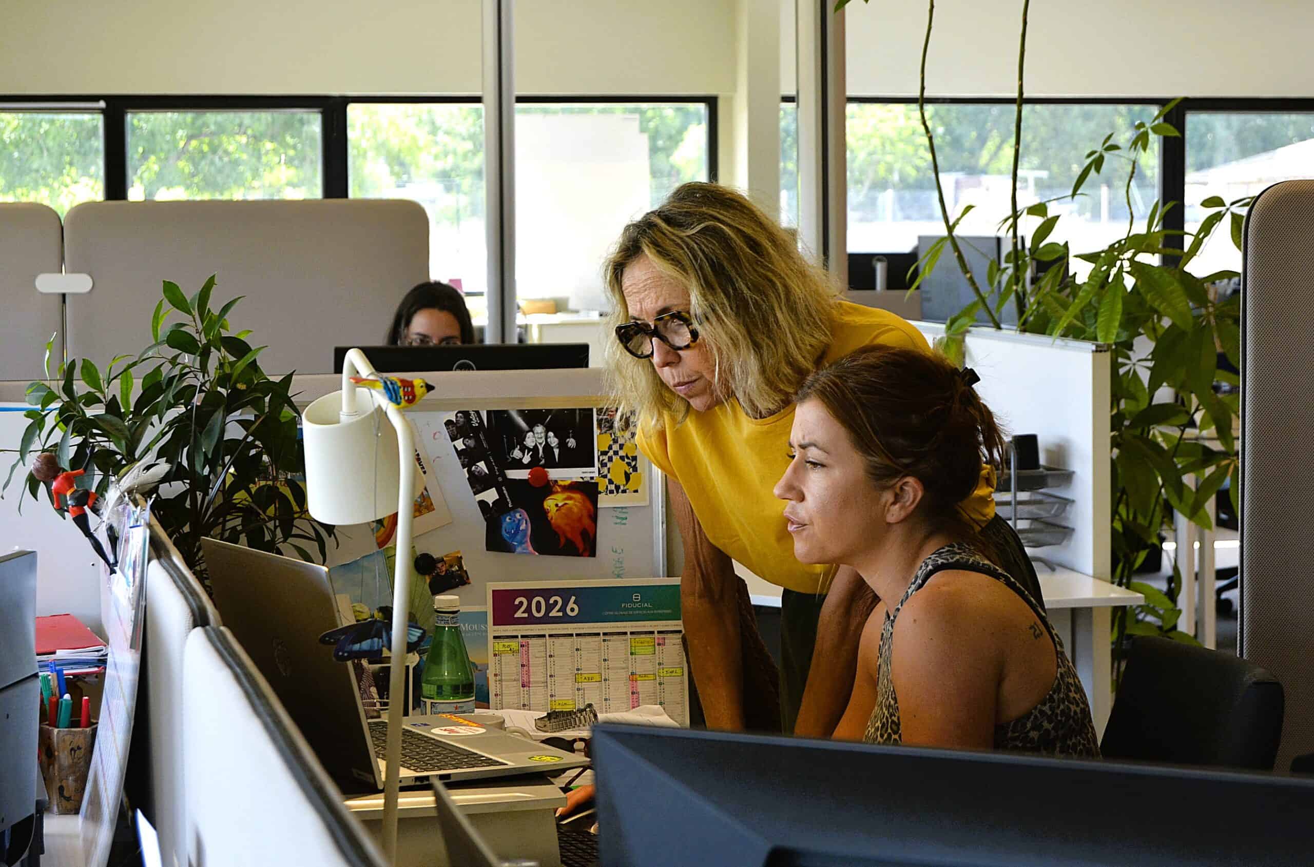 Two women collaborating at a modern office desk with plants and computers.