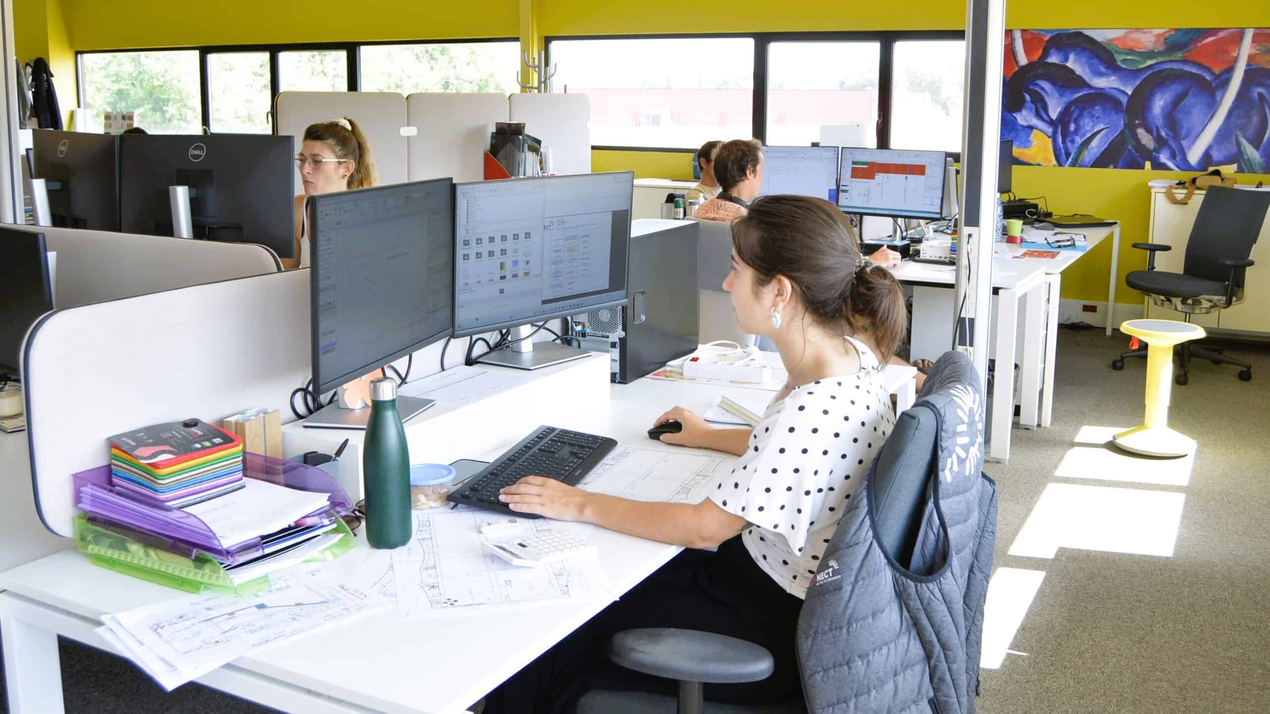 Modern office workspace with employees working at computers and dual monitors.